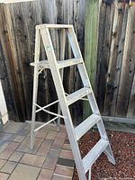 Full view of the 5ft aluminum step ladder standing on a tiled and gravel surface near a wooden fence.
