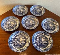 Seven blue and white shallow bowls arranged on a wooden table, showing scenic and floral blue designs.
