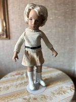 Blonde Sasha doll dressed in cream-colored outfit on white stand shown from front angle