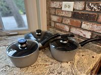 Set of three Berndes Germany non-stick cooking pots with glass lids and black handles, shown on a kitchen countertop with a brick backsplash.