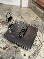 Brushed stainless steel napkin holder base showing tension arm mechanism and black clip on a granite countertop.