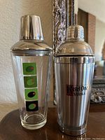 Pair of vintage martini shakers: one glass with avocado design, one stainless steel Ketel One Vodka shaker