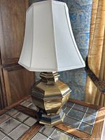Full view of vintage geometric brass table lamp with white fabric shade on a glass and wood side table.