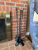 Photo of a vintage fireplace tool set including a poker, shovel, brush with dark wooden handles, standing on a metal tripod base in front of a brick fireplace.
