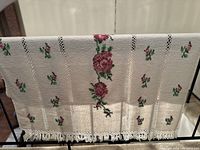 Full view of the white woven hand towel hanging over a rail, showing pink rose floral woven details and fringed edge.