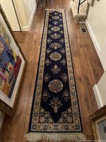 Photo of a navy blue long runner rug placed on wood floor, showing the entire length with repeating floral medallions and beige border with fringe ends.