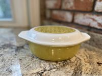Vintage 1970 Pyrex oval casserole dish in green color with its opal glass lid, shown against a kitchen countertop with brick behind.
