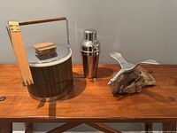 Three vintage items on wooden surface: wood and metal ice bucket with wooden handle, polished metal cocktail shaker, abstract aluminum sculpture on wood base