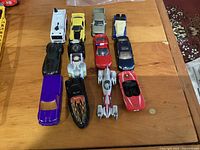 Twelve assorted toy vehicles arranged in three rows on a wooden surface, showing varied colors and designs including cars and a motorcycle.