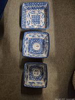Top-down view of the 3 stacked square pottery serving dishes with blue, white and orange floral decorative pattern, placed on carpet.