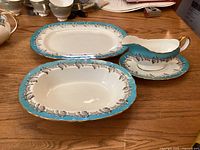 Oval platter, vegetable bowl, and gravy boat with underplate displayed on wood surface. Items feature turquoise border with oak leaf design and gold trim.