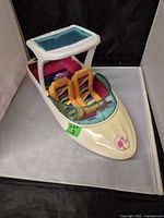 Front and top view of a white and pink Barbie toy speedboat with two yellow and purple striped seats and a blue windshield.