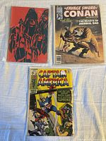 Lot of three comics displayed flat including a Sabretooth anthology with red graphic cover, Savage Sword of Conan #27 featuring Conan fighting a beast, and Captain America #123 showing Captain America in action.