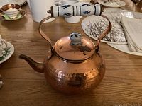 Copper kettle featuring a hammered texture, curved spout, ceramic knob and handle with blue decorative marks, placed on a wooden table.