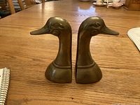 Pair of brass duck head bookends placed on wooden table, showing side profile of each head facing opposite directions.