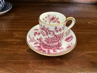 White ceramic espresso cup with red floral peony pattern and gold trim on wooden surface.