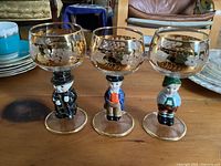 Front view of 3 Goebel Hummel wine glasses with figurine stems showing detailed painting and gold grapevine decoration