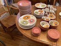 Full lot arranged on wood table includes stacks of dinner plates, lunch plates, cereal bowls, cups, saucers, creamers, and a covered vegetable bowl, showing overall colors and styles in the set.