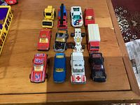 12 Majorette toy vehicles arranged in 3 rows of 4 on wooden surface; includes cars and trucks in various colors with playwear visible.