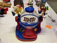 Front view showing clear globe candy container, red and blue base, and yellow and red M&M's character figurines attached on each side.