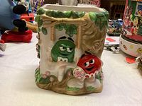 Front view of ceramic vase with green M&M character on tree branch and red M&M character holding flowers