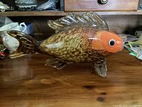 Side view of Murano glass fish figurine showing orange head, speckled body and clear glass fins and tail, displayed on wooden surface with background items.