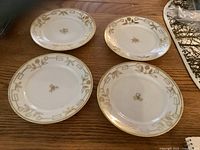 Four white porcelain bread plates with gold hand-painted floral and geometric pattern, arranged on wooden surface.