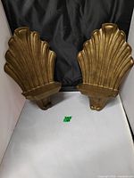 Front view of two gold-tone shell or fan shaped decorative wall shelves on white surface with black backdrop.