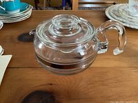 Clear side view of the Pyrex glass teapot showing handle, body, and lid on wooden surface with other plates in background.