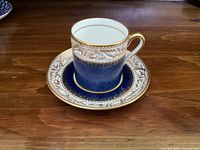 Blue and gold espresso cup and saucer set with intricate oak leaf and scroll gold pattern over a cobalt blue base, white cup interior, gold trim on cup rim and handle.