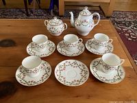 Full set arranged on wooden table showing teapot, sugar bowl, 5 cups, and 6 saucers all decorated with small pink flowers and green leaves pattern on white china.