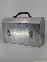 Front view of antique Aladdin tin lunchbox with embossed logo, black hard plastic handle, and metal clasps.