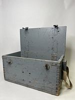 Photo of the open wooden storage box showing dovetail joints, canvas handles on sides, metal locking latches on front, and painted exterior with signs of wear.