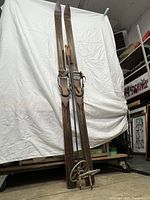 Full view of two antique wooden skis with bindings and poles in background, showing length and condition of wood and bindings