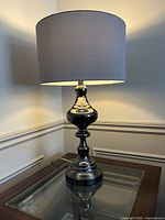 Black chrome table lamp on a glass-top side table, showing overall lamp and shade in natural lighting.