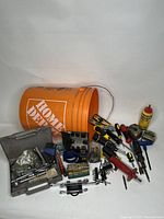 Wide shot of the Home Depot orange bucket lying on its side with various hand tools like screwdrivers, pliers, cutters, measuring tape, chalk line, and a bottle of lubricant visible outside the bucket.