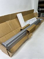 Aluminum finish exit device hardware partially out of its cardboard packaging on a white table.