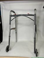 Front view of the silver folding walker showing dual 5-inch front wheels, adjustable legs, red folding release buttons, and soft handle grips.