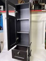 Black wooden storage cabinet standing upright, door open showing two shelves and bottom drawer open.
