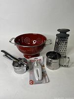 Full view of the kitchen accessory lot showing all five items arranged on a white background: Paderno strainer, potato ricer, flower sifter, box grater, and Starfrit can opener in packaging.