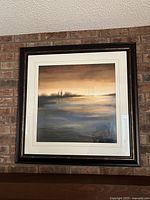 Framed large square sunset landscape art print hanging on brick wall, showing entire piece and frame.