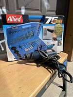 Photo of Allied 75-piece home/shop bit set in original box with blue plastic organizer case and Ryobi corded drill on wooden table.