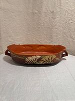 Front view showing the orange/brown colored oval ceramic bowl with raised floral decoration and double handles.