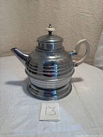 Front angled view of the chrome Art Deco teapot showing tiered raised ridges, Bakelite handle, and lid knob.