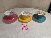 Three vintage Aynsley teacups and saucers displayed on a white cloth tablecloth, showing the pink, yellow, and turquoise blue cups and matching saucers lined up front-facing.