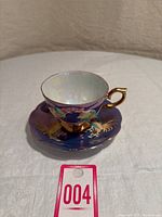 Side view of the teacup sitting on the saucer, showing the rich blue luster glaze and detailed raised rooster designs with gold handle and trim.