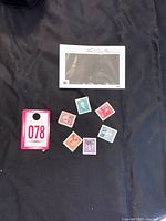 Top view of 6 Canadian Prime Ministers postage stamps arranged in a circle on black fabric along with lot number tag and a transparent plastic sleeve.