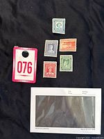 Photo showing six mixed mint and used Newfoundland postage stamps laid out on a black surface with a red and white tag labeled 076 and a plastic sleeve envelope.