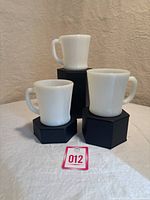 Three white milk glass coffee mugs with D handles displayed on black stands in varying heights, showing overall set and condition.