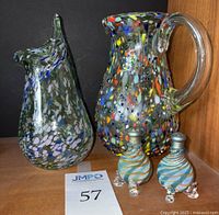 Four art glass pieces shown from front side on wooden surface against black background: confetti glass pitcher, green glass vase with blue and lavender accents, two blue swirl salt and pepper shakers with metal tops.